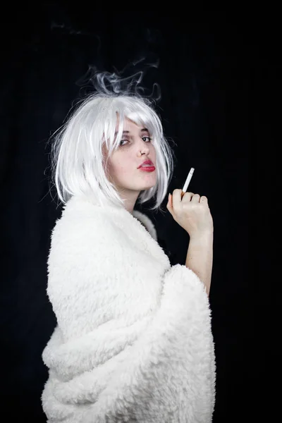 Attractive young woman in costume smoking cigar and looking at camera while standing on black background