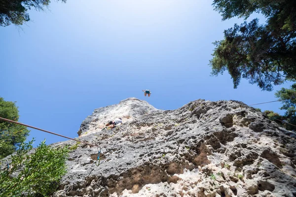 From below shot of climber secured with rope jumping from top of mountain under blue sky while other is climbing