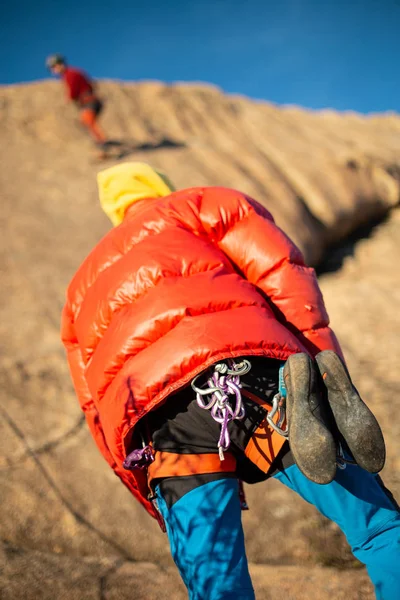 From below shot of man in warm coat looking up and checking climbing partner on high cliff in sunlight