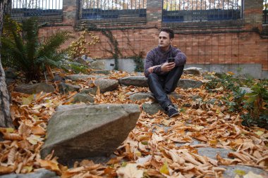 Genç yakışıklı bir rock ile yeşillik çayır üzerinde oturan ve smartphone ile konuşan adam