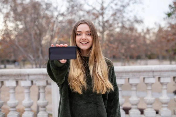 Lovely young female in stylish outfit smiling and looking at camera while standing in park and demonstrating modern smartphone with blank screen