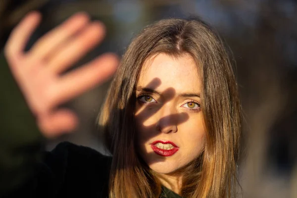 可爱的年轻女性与明亮的化妆试图覆盖脸与手从阳光 而站在模糊的背景公园 — 图库照片