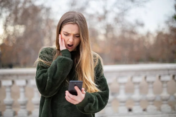 Troubled young woman rubbing forehead and looking at modern smartphone while standing in autumn park
