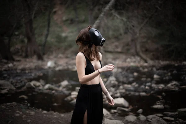 Portrait of anonymous woman in black clothes and gas mask standing in amazing spooky forest