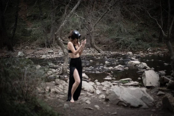 匿名赤脚妇女穿着黑色衣服和防毒面具站在惊人的怪异森林侧视图 — 图库照片