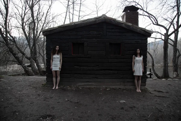 身着白衣的孪生姐妹们站在神秘森林中废弃的木屋旁 — 图库照片