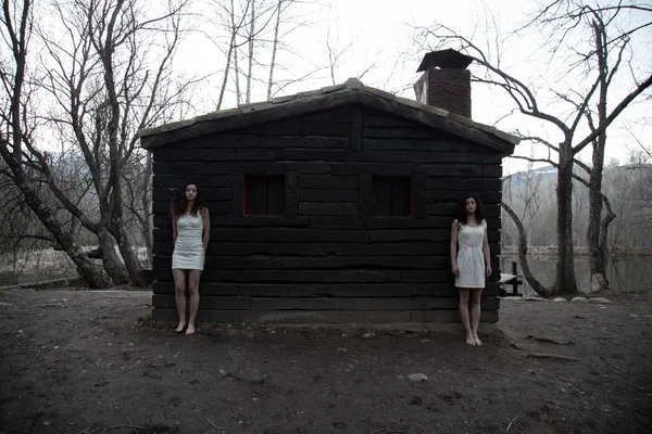 身着白衣的孪生姐妹们站在神秘森林中废弃的木屋旁 — 图库照片
