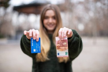 Genç bayan parkta dururken kameraya plastik kart ve para gösteriyor.
