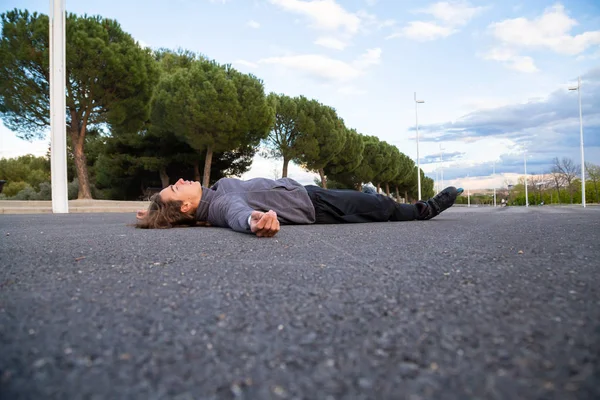 Young fit weary man in sportswear with roller skates lying on road in beautiful city park