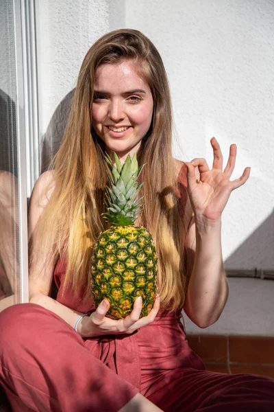 Young cheerful sensual woman in casual summer dress holding tropical pineapple with closed eyes and showing ok gesture