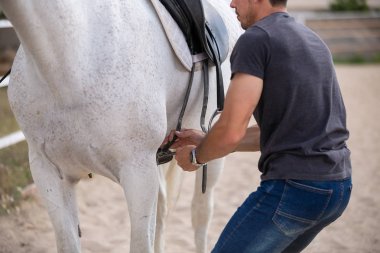 Çiftlikte ki padokta dururken beyaz atın arkasına siyah eyer koyan genç adamın yan görünümü