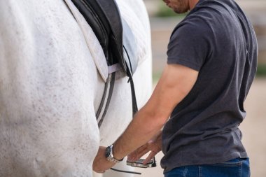 Çiftlikte ki padokta dururken beyaz atın arkasına siyah eyer koyan genç adamın yan görünümü