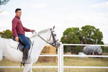 Çiftlikte kumlu zeminde beyaz ata binen günlük kıyafetli genç adam.