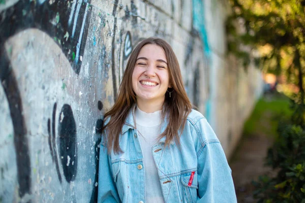 Pretty young female in casual outfit cheerfully smiling and leaning on weathered graffiti wall while standing on city street on sunny day