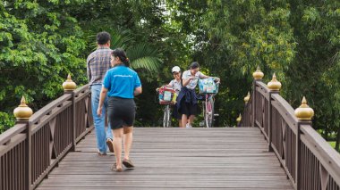 Bangkok, Tayland - 25 Eylül 2018: Bisikletli gülen Taylandlı kızlar Bang Krachao 'daki bir parkta tahta bir köprüden diğer yolculara doğru yürüyorlar.).