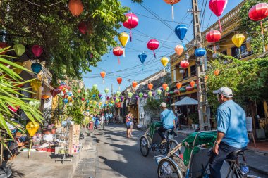 Hoi An, Vietnam - 24 Ekim 2018: İki triportör, fenerlerle süslenmiş eski şehrin renkli bir caddesi boyunca ilerliyor..