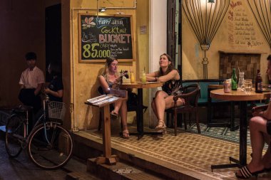 Hoi An, Vietnam - 13 Kasım 2018: İki genç kadın gezgin eski kasabadaki bir barda oturuyorlar, sokaktan bir manzara.