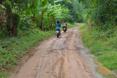 Nam Tok Sai Yok Noi, Tayland - 18 Ağustos 2018: Üzerinde üç kişi bulunan iki scooter toprak yol boyunca (Sai Yok Noi şelalesi üzerinde) tropikal bitki örtüsünün yeşilliğine doğru yola çıkıyor.