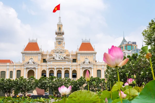 Saigon City Hall (or Ho Chi Minh City People\'s Committee) with pink lotus flowers and blooming plumeria trees in the foreground (blurred). One of the top tourist attractions of the city. Vietnam 2019.