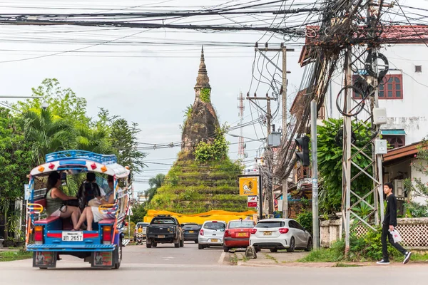 Vientiane, Laos - 9 Ağustos 2018: Dam Stupa çimen ve çalılarla kaplandı, insanlarla dolu bir tuk-tuk, yayalar, kalın pleksus elektrik kabloları ve sokak bitkileri de dahil olmak üzere sokak trafiği.