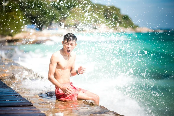 Young Asian man sits on a pier with the excited face & expressive hand gesture being wet under the shower of sea spray after a large wave hit on a sunny day on Koh Samet, Thailand. Soft focus.