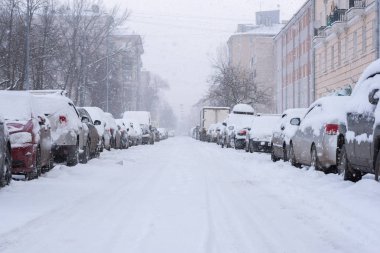 Boş sokakta kar yağışı. Karla kaplı sıra sıra park edilmiş arabalar. Düşen kar tanelerinin arasından görünen binalar ve parklar. Şehirde beyaz kış. Opochinina Caddesi, Vasilyevsky Adası, St. Petersburg.