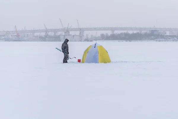 Saint Petersburg, Rusya - 4 Şubat 2018: balıkçı, donmuş Neva Nehri 'ndeki bir buz balıkçı çadırının yanında bir buz matkabı tutuyor. Çadırın içinde iki balıkçı daha var. Buz balıkçılığı Ruslar arasında popüler bir hobidir..