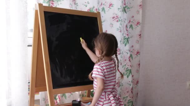 Petite fille d'âge préscolaire écrivant des lettres sur tableau noir. Petite fille tenant craie et dessin . — Video