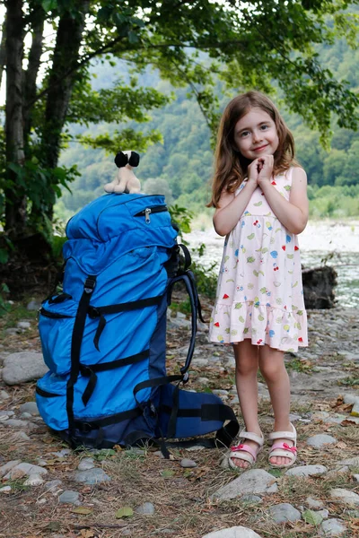 A large backpack for an adult stands next to a little girl against the backdrop of trees and a river, on a sunny day. — Stock Photo, Image