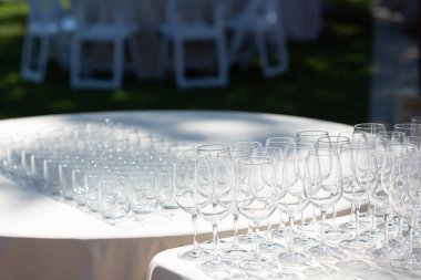 Sıralar dolusu parıldayan boş şarap kadehleri ve bardaklar bir açık hava etkinliğinde beyaz masa örtüleriyle masalarda hazır bekliyor, misafirleri ve içecekleri bekliyor.