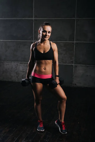 Young woman bodybuilder with perfect athletic body is posing with two dumbbells