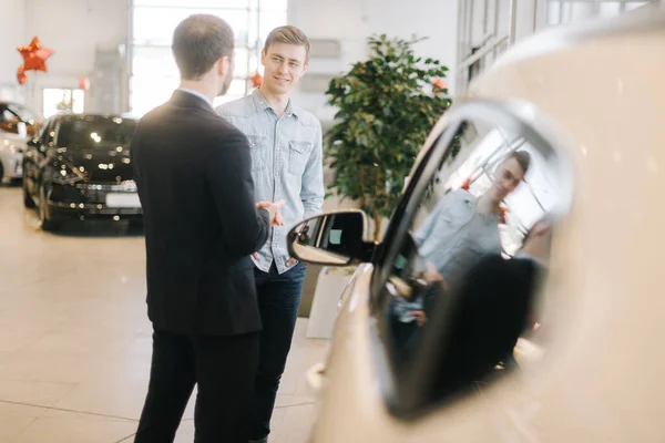 Confident car salesman wearing business suit is telling about new car model to young man.