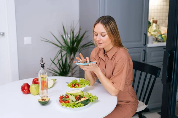 Happy young vegetarian woman uses smartphone for taking photos of vegetables.