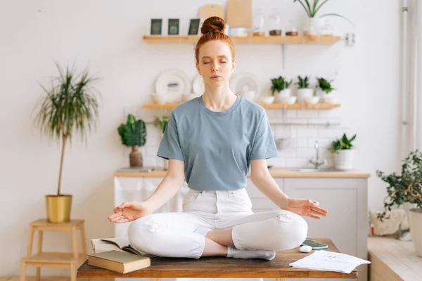 Peaceful redhead young woman is sitting with closed eyes on table in lotus position