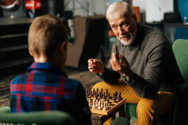 Mature adult grandfather explaining rules of chess game to grandchild at home in cozy living room.