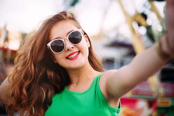 カルーセルの前で Selfie を作ってピンクのサングラスのうれしそうな笑顔の女の子 遊園地で楽しくかわいいスタイリッシュな女性の屋外のポートレート — ストック写真