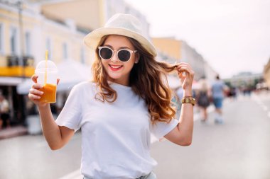 Parlak makyajlı, enerjik harika bir modelin portresi. Kısa kot şortlu, parlak tişörtlü, güneş gözlüklü, lunaparkta şenlik yapan sevimli bir kız. kokteyl içerir