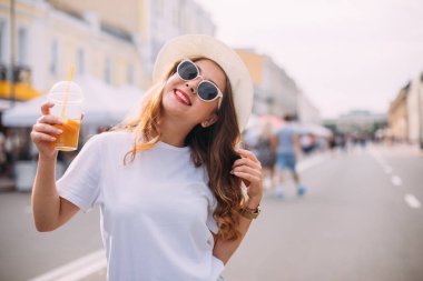 Parlak makyajlı, enerjik harika bir modelin portresi. Kısa kot şortlu, parlak tişörtlü, güneş gözlüklü, lunaparkta şenlik yapan sevimli bir kız. kokteyl içerir