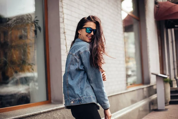 Joyeux attrayant jeune femme en lunettes de soleil marche dans la ville. Femme ludique s'enveloppe par-dessus son épaule et regardant dans la caméra . — Photo