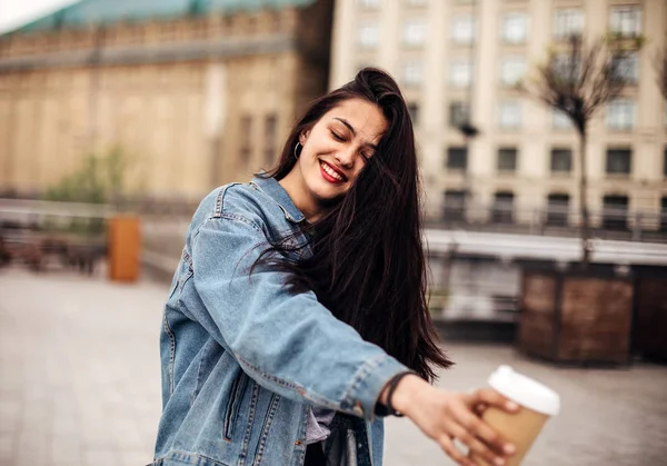 Utomhusporträtt av en mörkhårig ung kvinna i jeansjacka håller kaffet uppe. Känslomässig kvinna poserar med glada leenden på gatan. — Stockfoto