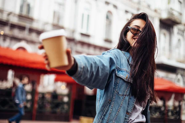 Stylish happy young woman wearing woman in a denim jacket. She holds coffee to go. portrait of smiling girl in sunglasses and bag — Stock Photo, Image