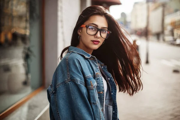 Närbild porträtt av bedårande flicka poserar på gatan. flicka student i en denim jacka och glasögon tittar i kameran — Stockfoto