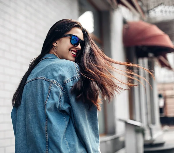 Joven modelo hermosa chica en gafas de sol sonríe y mira hacia atrás en la ciudad. Dinámicamente chica joven camina por la calle. Cabello revoloteando en el viento . —  Fotos de Stock