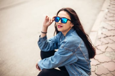 Genç bir kız öğrenci kaldırımda oturuyor. Kot pantolon giy ve kahve iç.