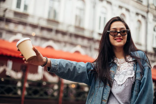 Snygg glad ung kvinna klädd kvinna i jeansjacka. Hon har kaffe att ta med. Porträtt av leende flicka i solglasögon och väska. Känslomässig kvinna — Stockfoto