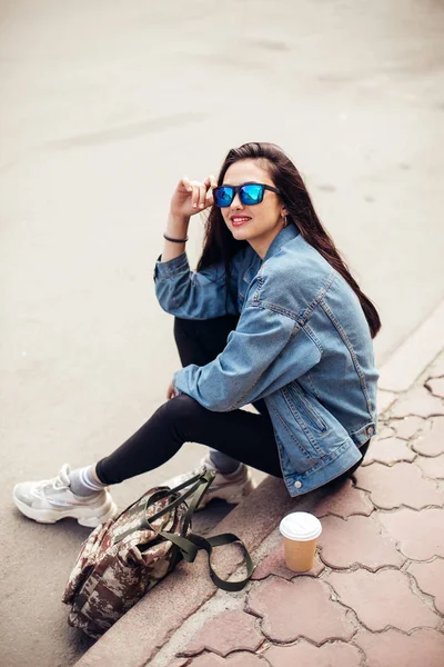 Ung flicka student sitter på trottoaren. Bär jeans och drick kaffe. Ung vacker modell flicka i solglasögon leenden — Stockfoto
