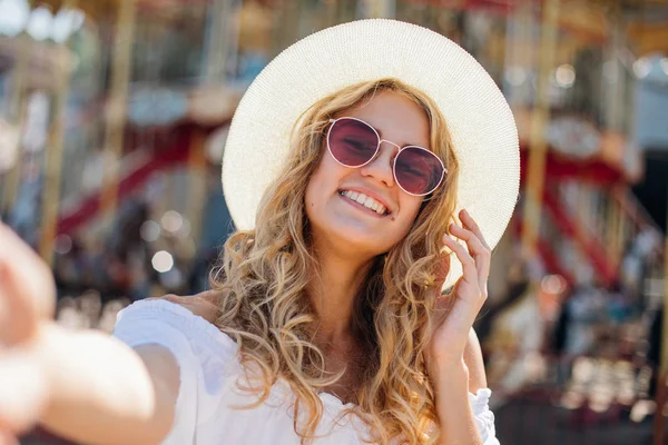 Chica sonriente alegre en gafas de sol de color rosa haciendo selfie delante del carrusel. Retrato al aire libre de mujer elegante lindo divertirse en el parque de atracciones y tomar selfie —  Fotos de Stock