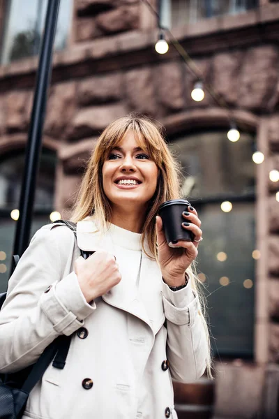 Stylish happy young woman wearing beige trench coat.She holds coffee to go. portrait of smiling girl . Street fashion concept — Stock Photo, Image