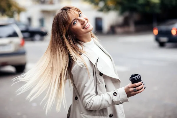 Stylish beautiful woman blonde in beige trench coat.She holds coffee to go. portrait of smiling girl . Street fashion concept — Stock Photo, Image