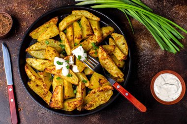 Bir tavada, yeşil soğan, baharat sıcak patates kızartması. Beyaz sos. Patates bir rustik tarzda pişirilir. Koyu arka plan, çatal, bıçak.